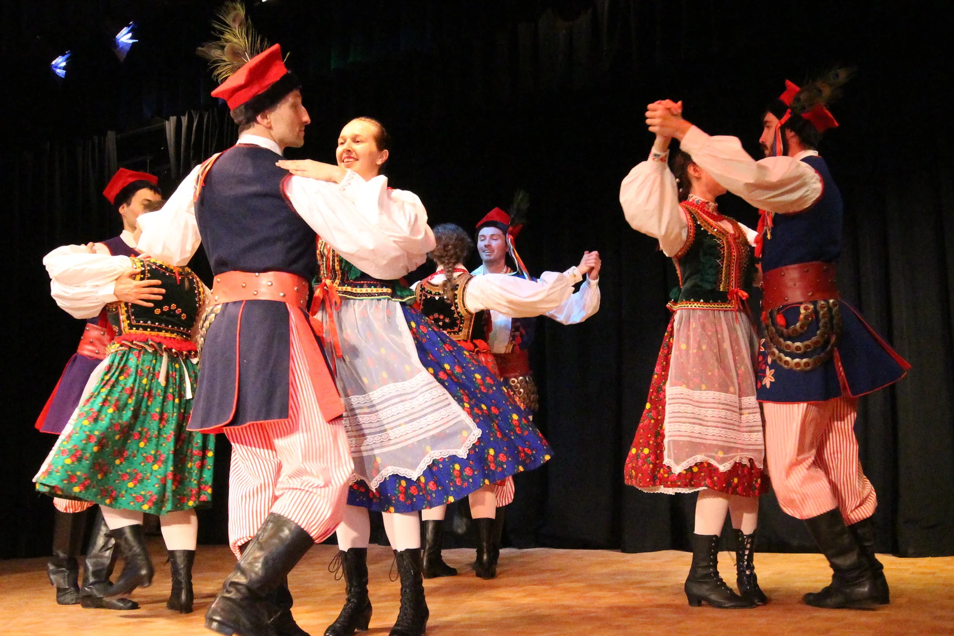 Ballerini in costume tradizionale di Cracovia — copricapi con piume di pavone, grembiuli rossi e stivali neri — mentre danzano su un palco illuminato