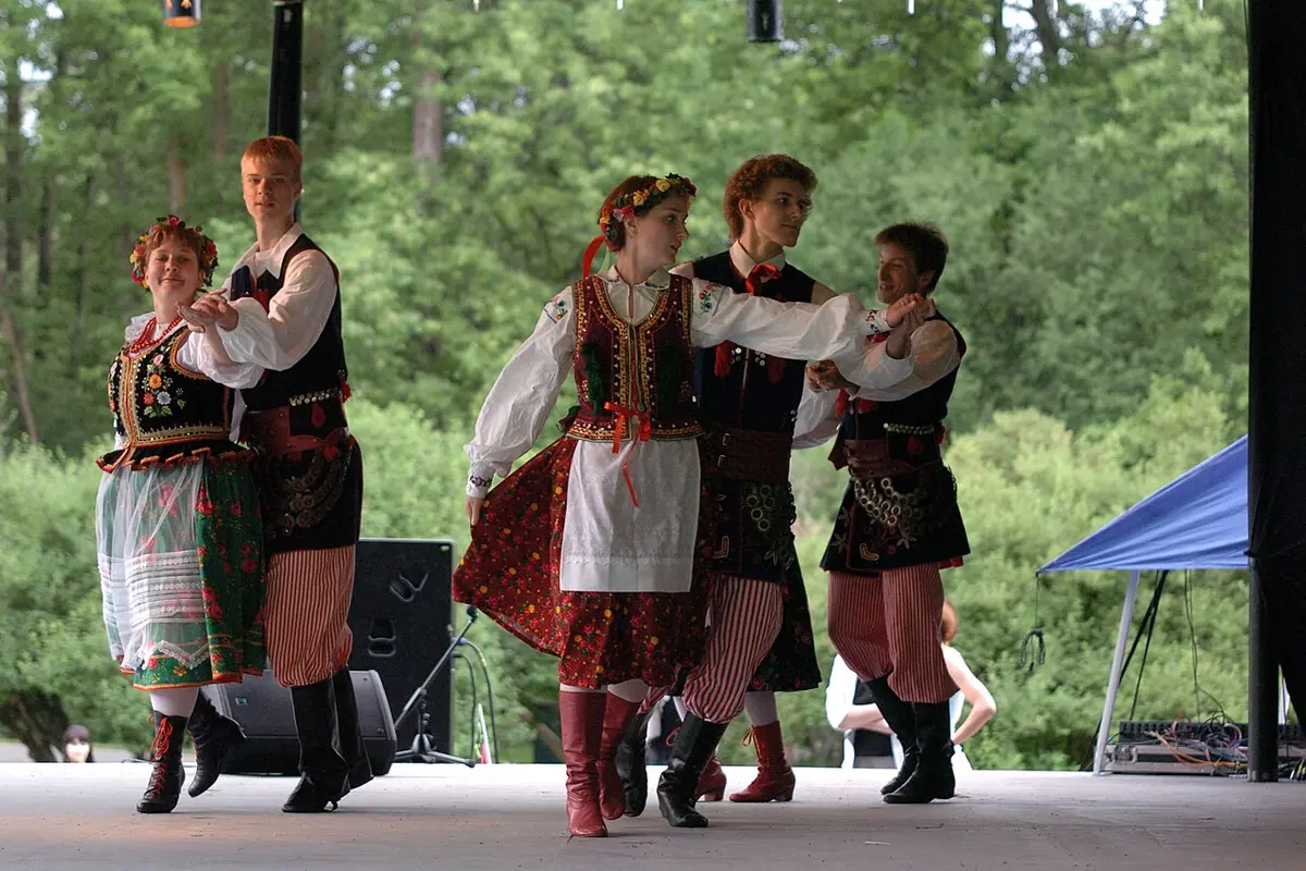 Four dancers in Polish folk costume with flower wreaths, embroidered vests, striped trousers and tall boots, dancing on a stage in front of trees