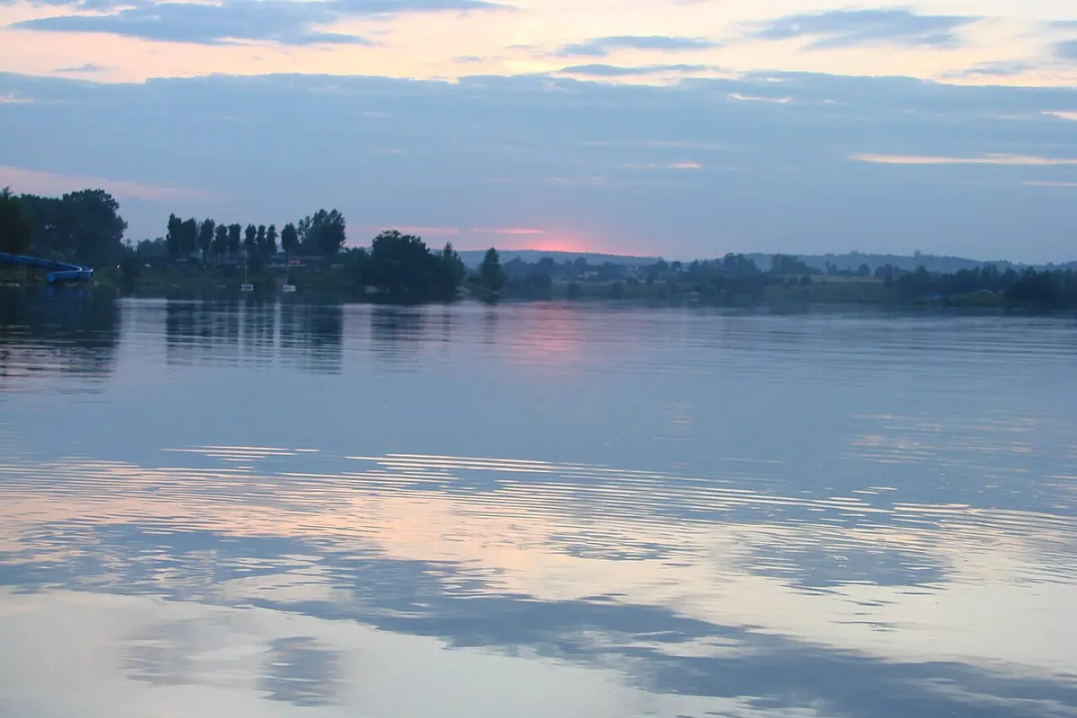 A calm reservoir at dusk with a pink and blue sky, tree-lined shores and a blue water slide at the far left